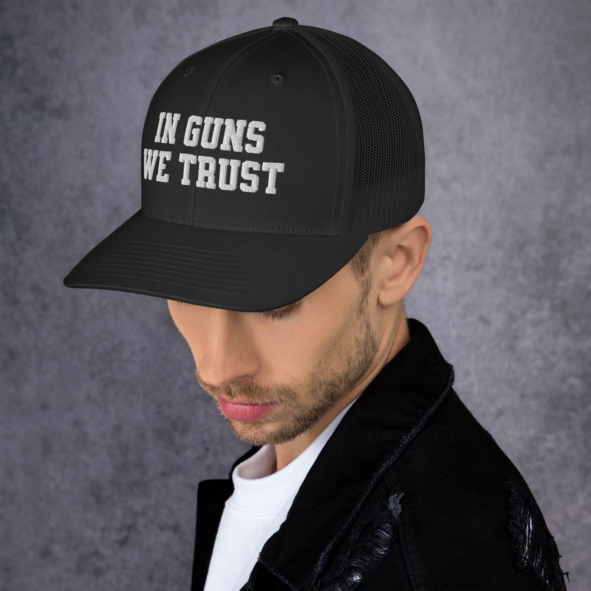 Man wearing a black cap with 'IN GUNS WE TRUST' text on a gray background