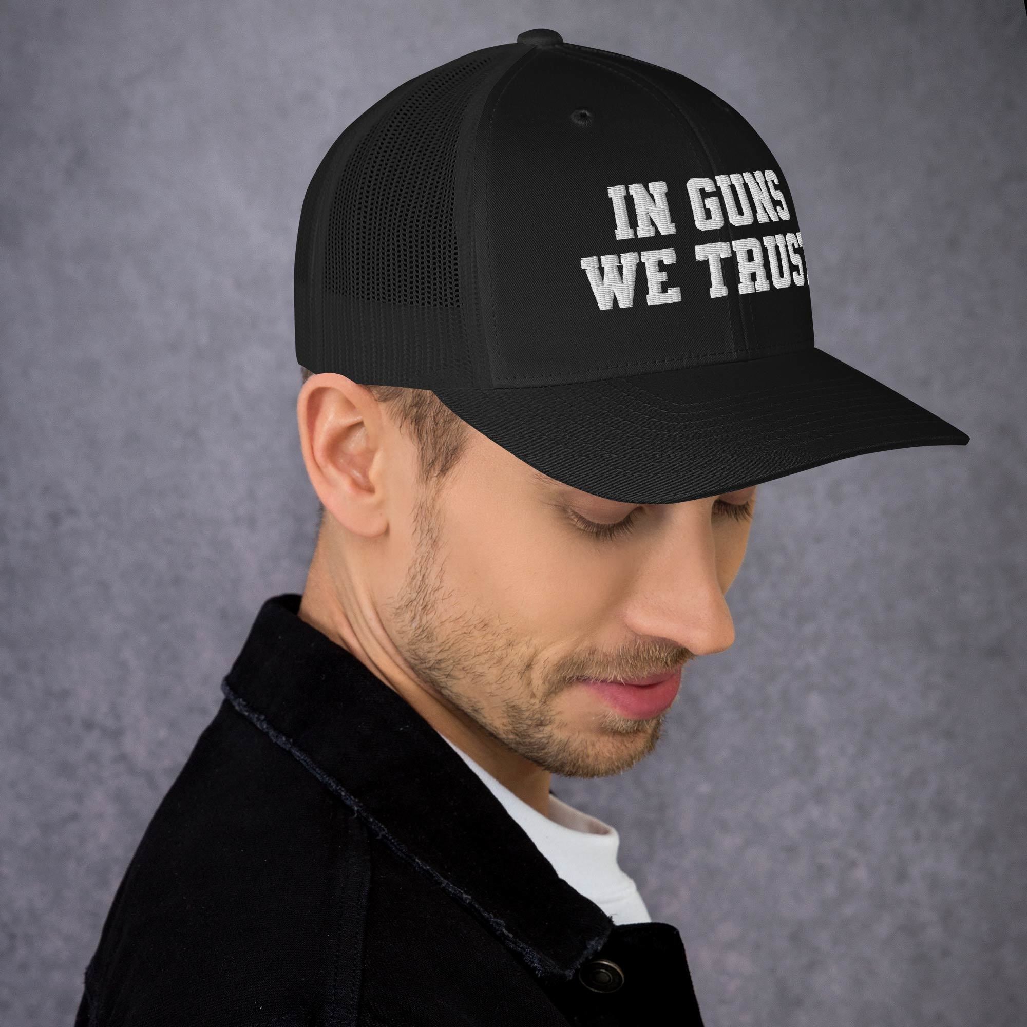 Man wearing a black cap with 'IN GUNS WE TRUST' text on a gray background