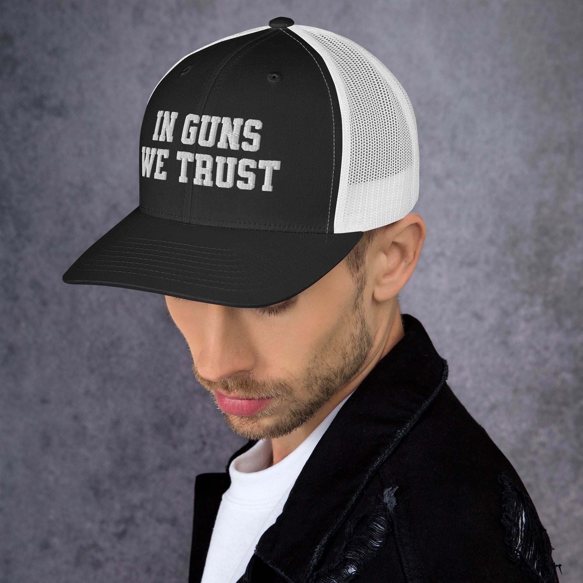 Man wearing a black and white cap with 'IN GUNS WE TRUST' text on a gray background