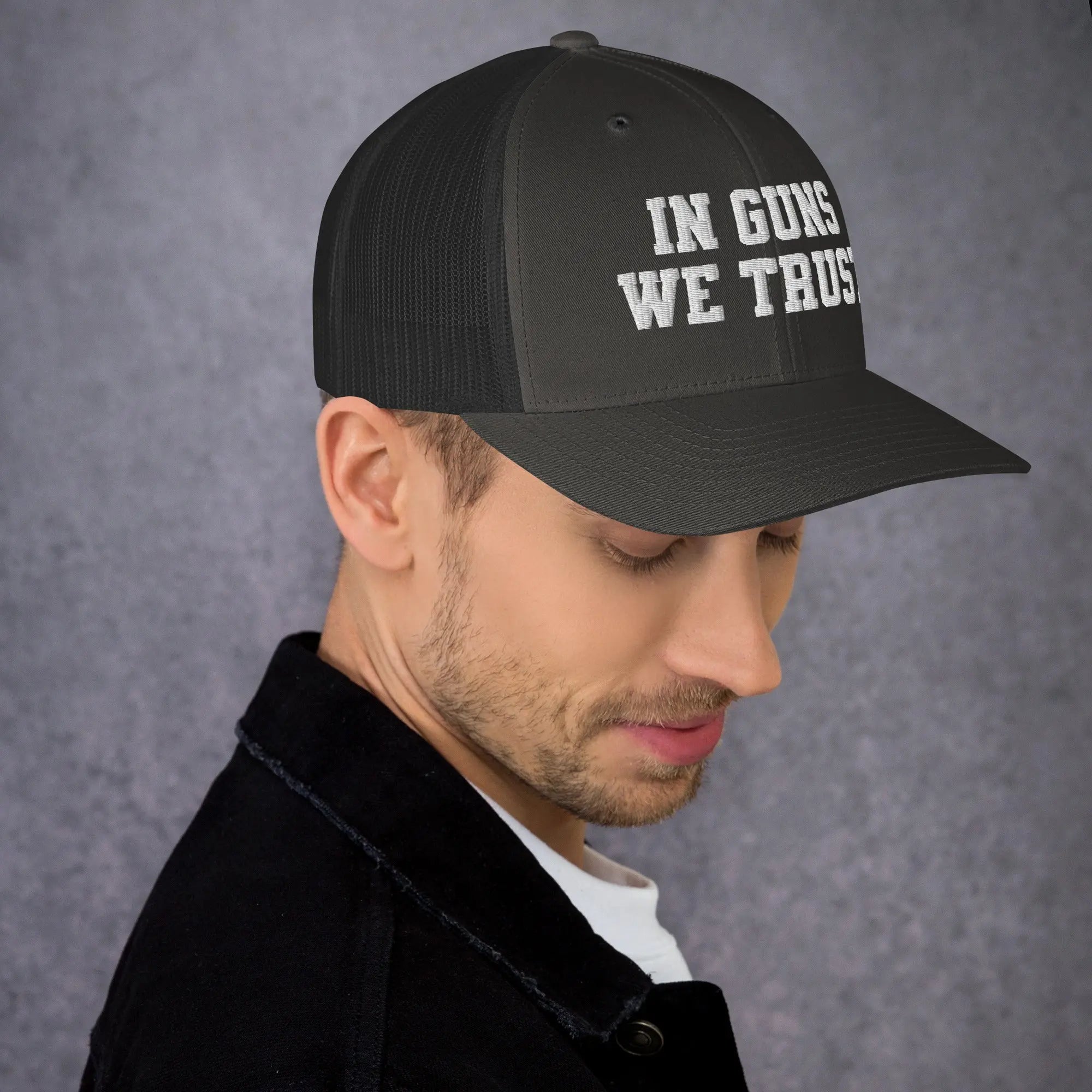 Man wearing a black cap with 'IN GUNS WE TRUST' text on a gray background