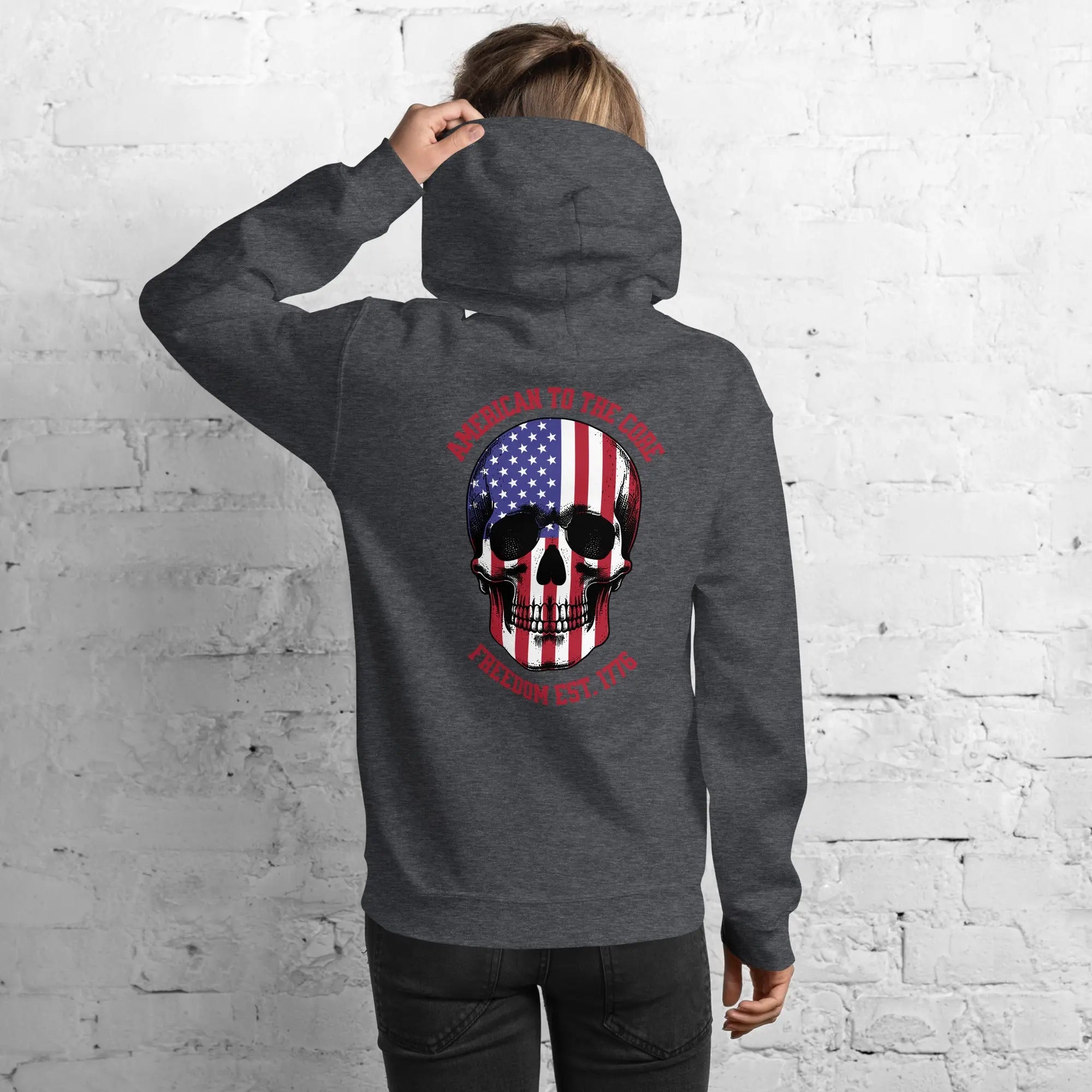 Back of a woman wearing a dark heather patriotic hoodie with a skull and the text "American to the core. Freedom Est. 1776." 