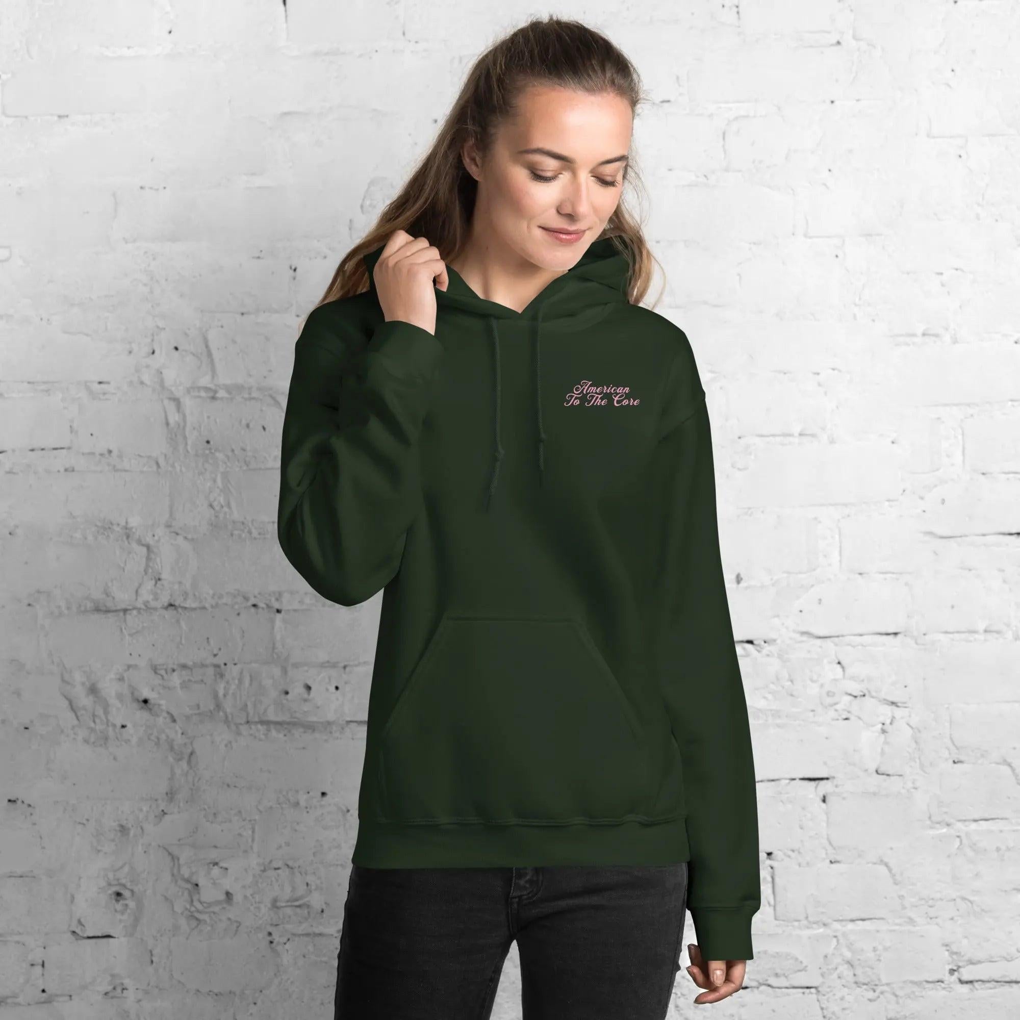 A woman wearing a forest green patriotic hoodie with the text "American To The Core"