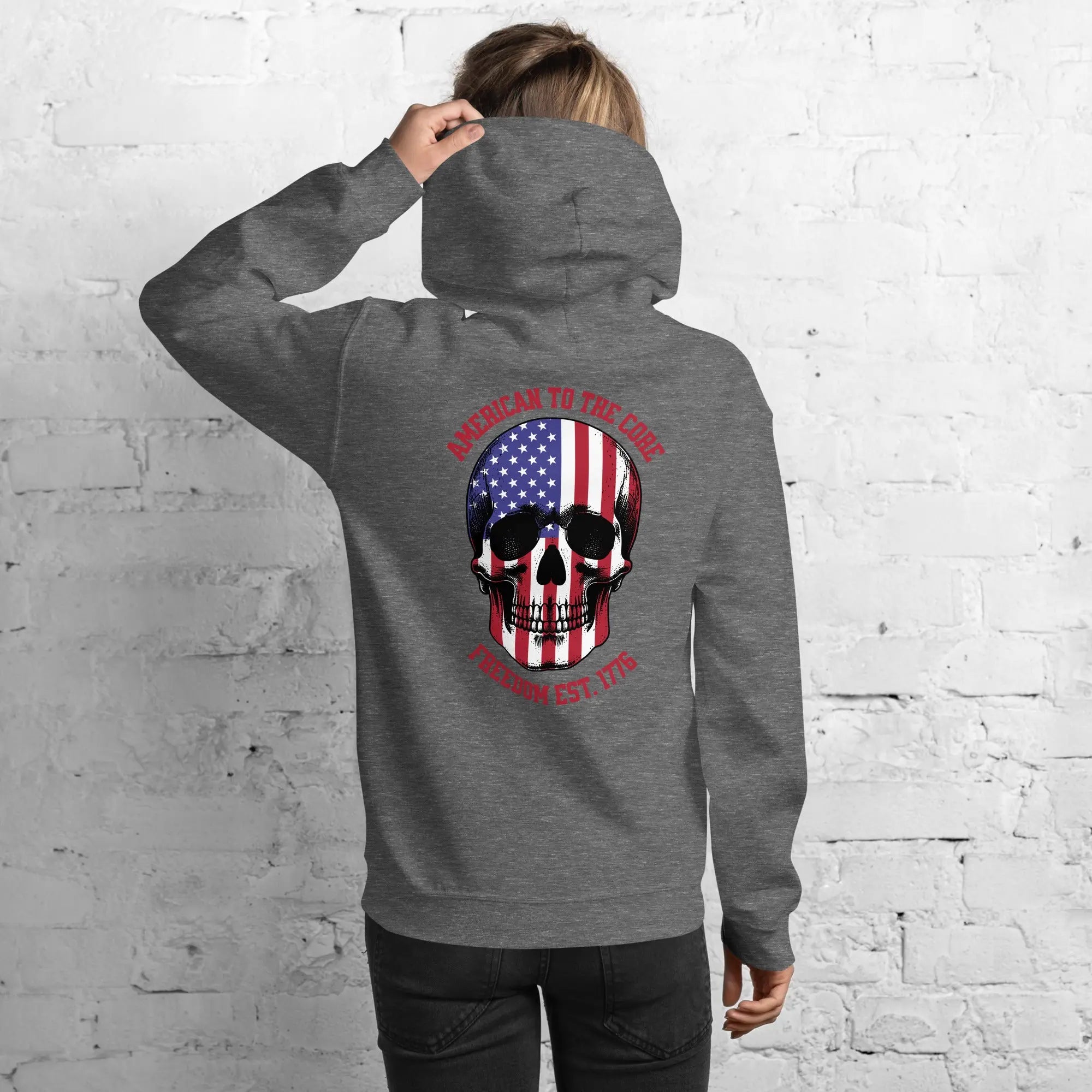 Back of a woman wearing a graphite heather patriotic hoodie with a skull and the text "American to the core. Freedom Est. 1776." 