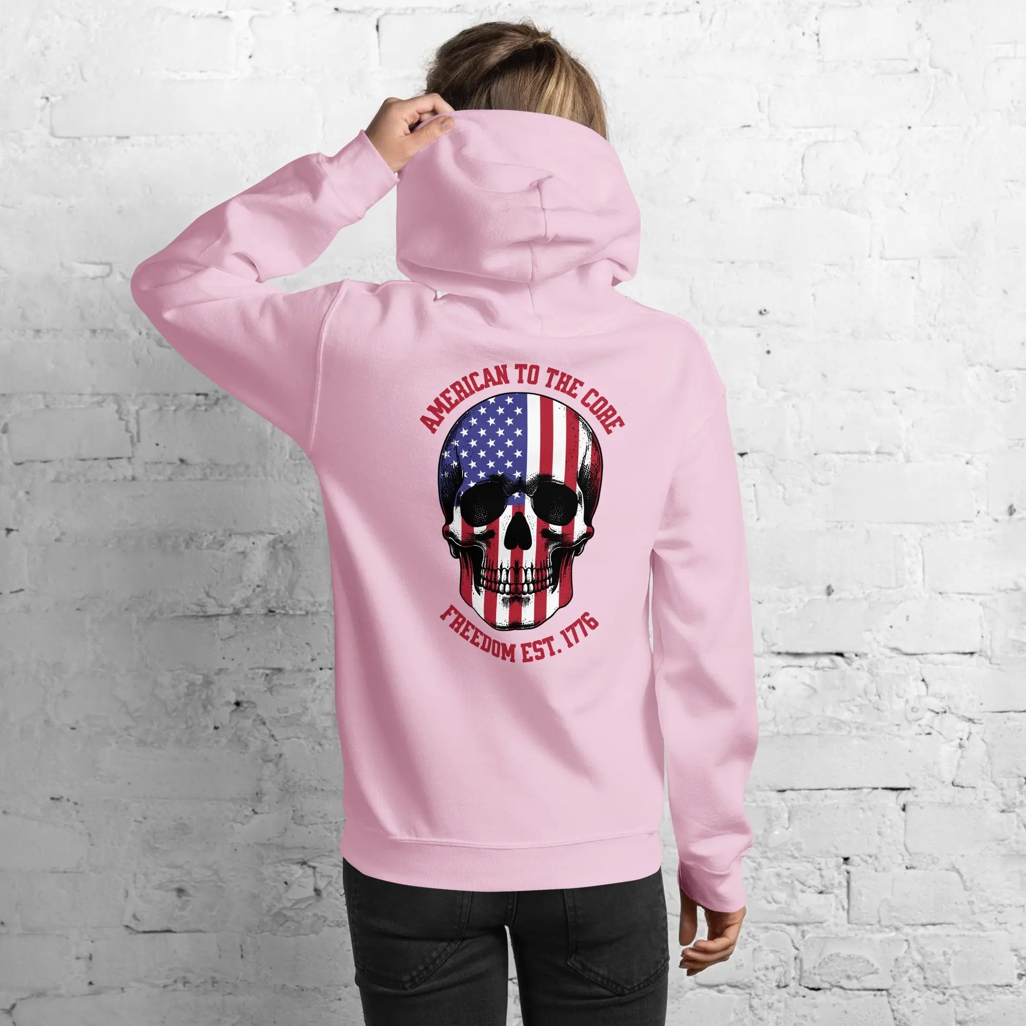Back of a woman wearing a light pink, patriotic hoodie with a skull and the text "American to the core. Freedom est. 1776." 