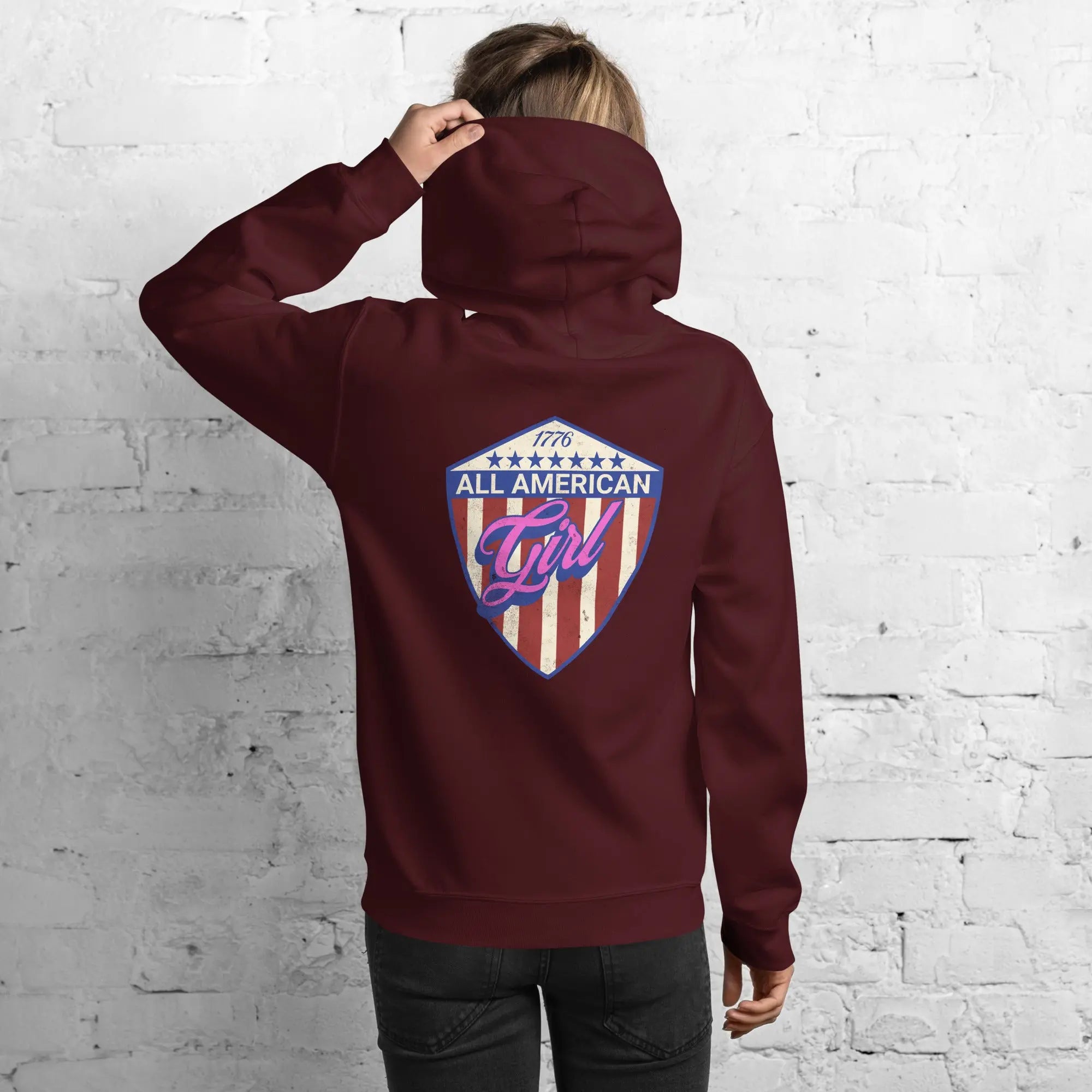 Back of a woman wearing a patriotic maroon hoodie with the text "All American Girl" over a patriotic design. 