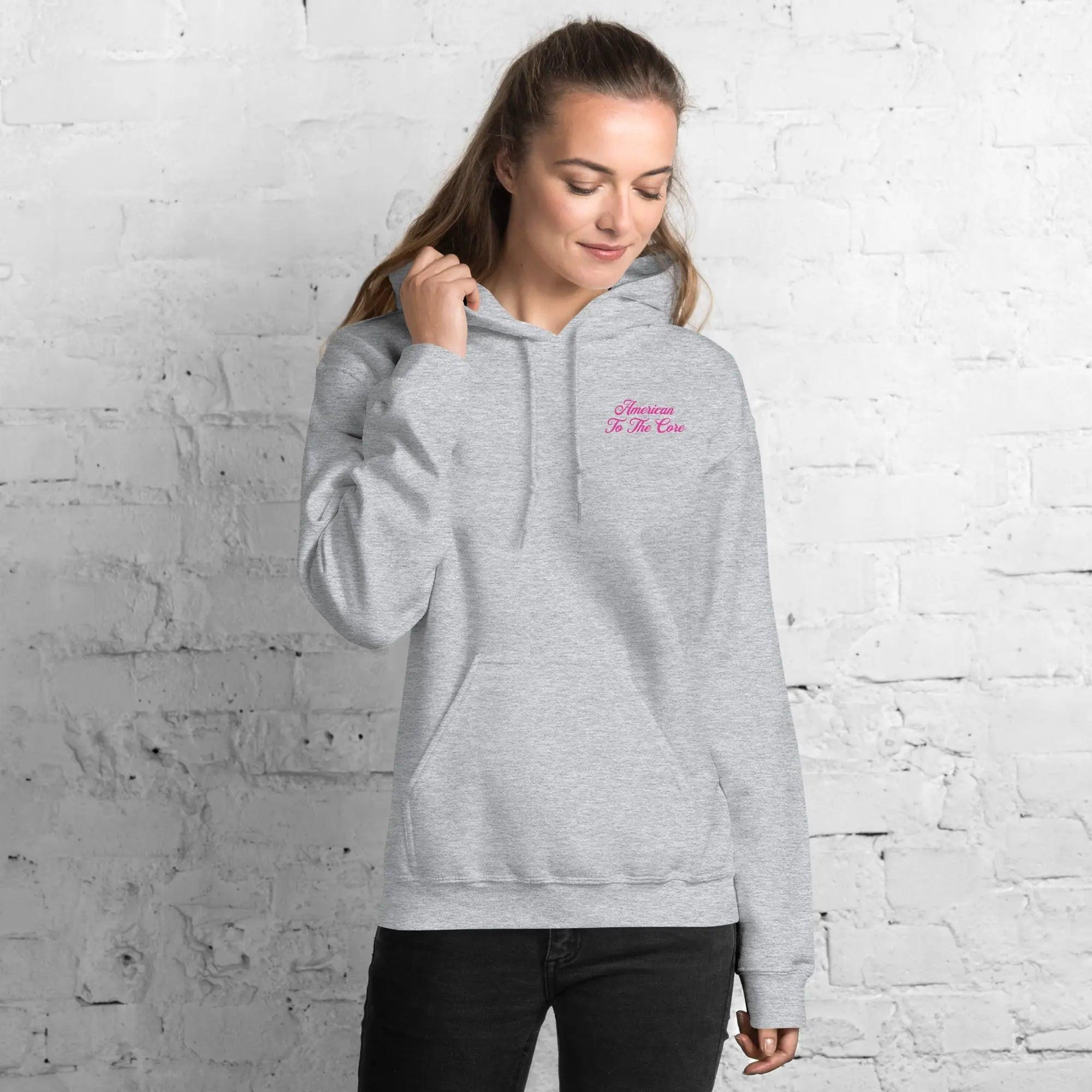 A woman wearing a patriotic sport grey hoodie with the text "American To The Core" in pink.