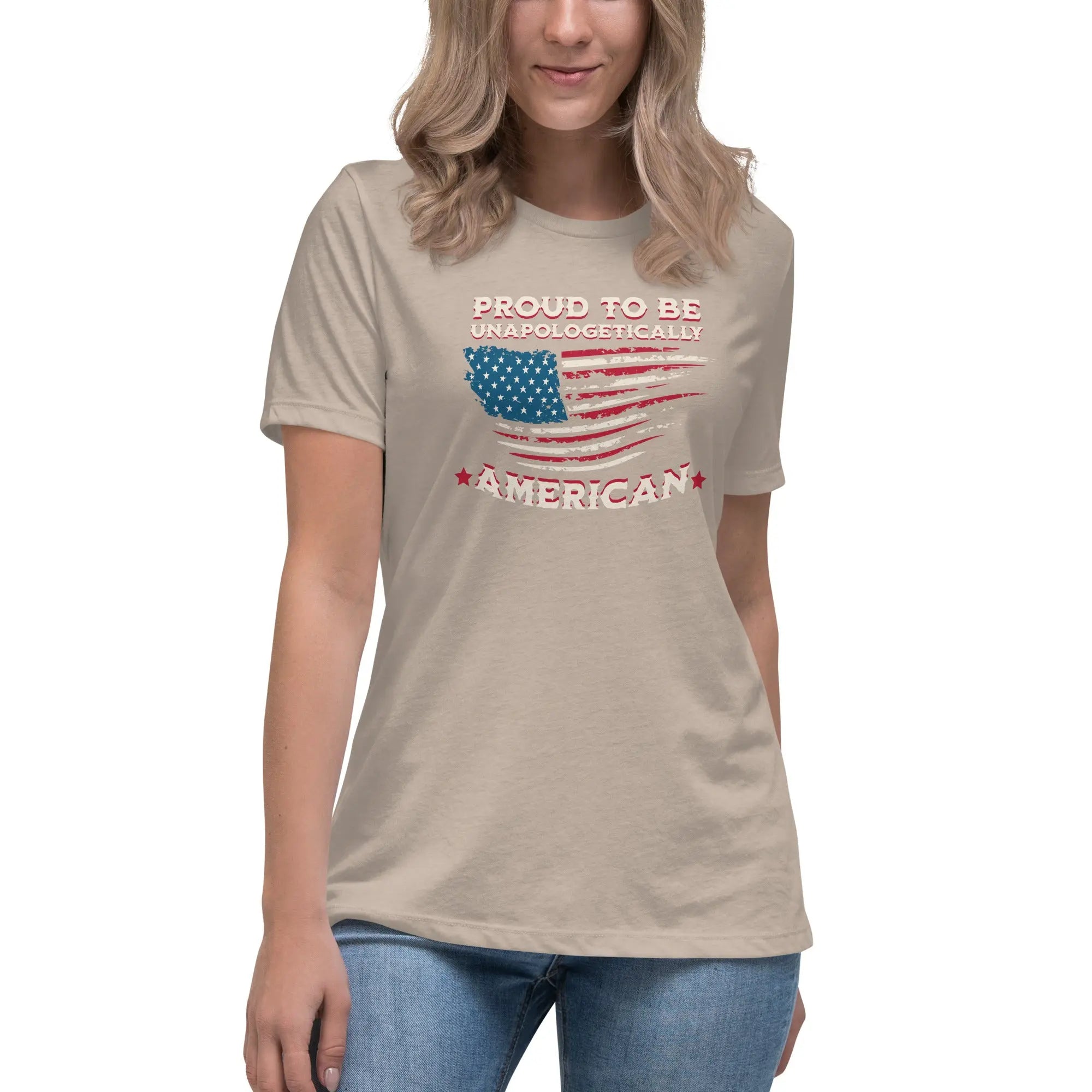A woman wearing a heather stone patriotic t-shirt with the American flag and "Proud To Be Unapologetically American" text.